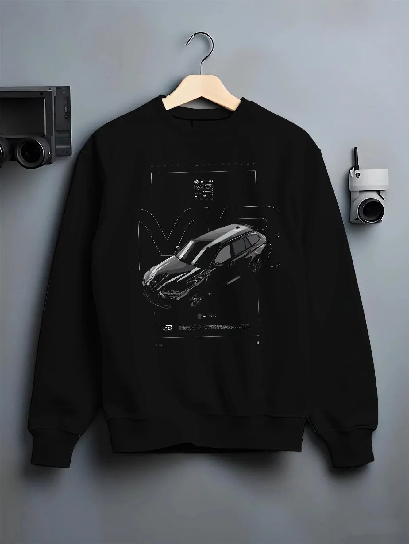 Black crewneck sweatshirt on a hanger against a grey wall, featuring BMW M3 street racing performance motorsport automotive graphic.