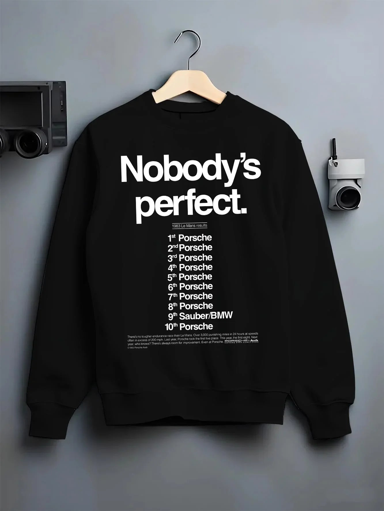 Black crewneck sweatshirt on a hanger against a grey wall, featuring Porsche rs racing le mans motorsport street graphic.